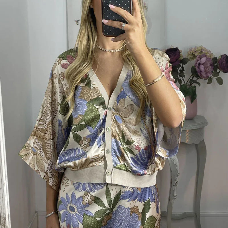 A woman is taking a mirror selfie wearing a satin V-neck floral blouse with a matching ankle-length skirt with a neutral background.