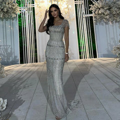 Woman in a silver evening gown standing outdoors with decorative flowers and white railing in the background.