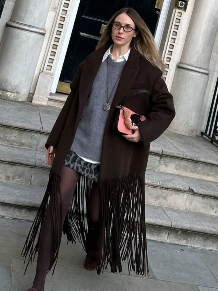 Street-style shot of a woman wearing a brown fringed trench coat over layered knits, standing on stone steps outside a building.