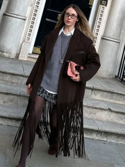 Street-style shot of a woman wearing a brown fringed trench coat over layered knits, standing on stone steps outside a building.