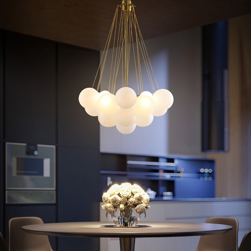 A frosted glass chandelier with a silver iron finish hangs over a round dining table. The chandelier has a white color, and it appears to be dimly lit, enhancing the cozy ambiance of the room.