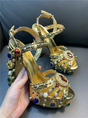 Gold high-heeled shoes with gemstones held by a hand against a dark background
