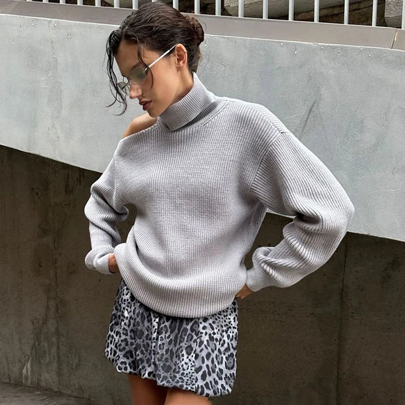 Woman wearing a gray turtleneck off-shoulder loose knit sweater styled for autumn and leopard print skirt against a concrete wall.