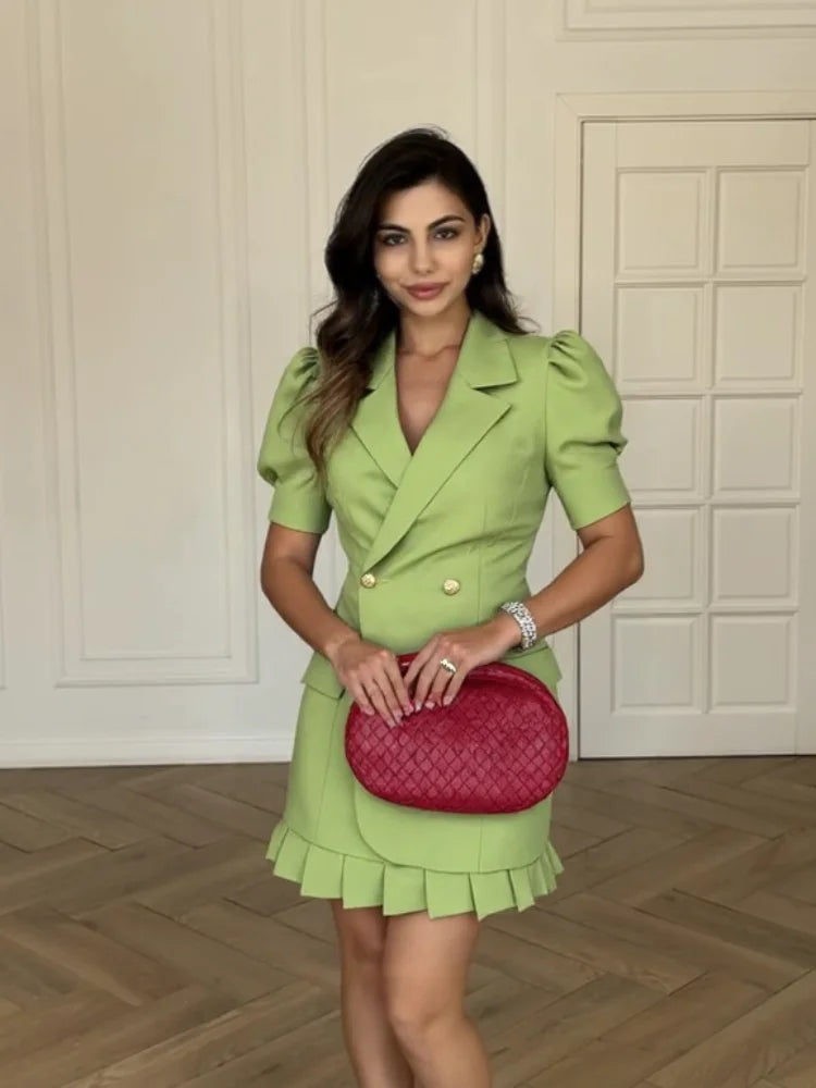Woman in a Green pleated skirt and blazer set styled with a red bag against a white panelled wall.