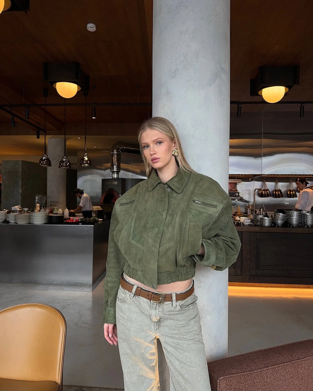 Woman in a green suede zip bomber jacket styled for autumn and winter streetwear, standing in a modern interior setting