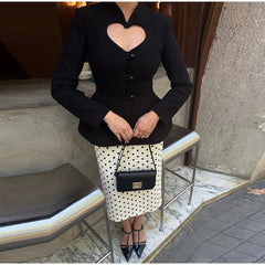 Woman wearing a black short blazer with a heart cutout and button front, styled with a polka dot midi skirt and pointed heels outdoors.