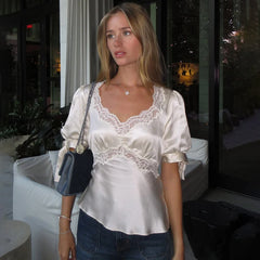 Model wearing an ivory satin-finish top with lace-trim V-neck, styled with denim and a shoulder bag in evening lighting.