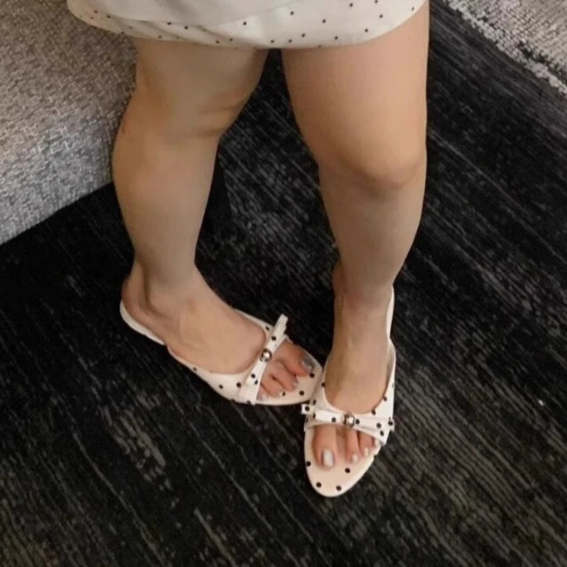 Person wearing white sandals with black polka dots on a dark textured floor.