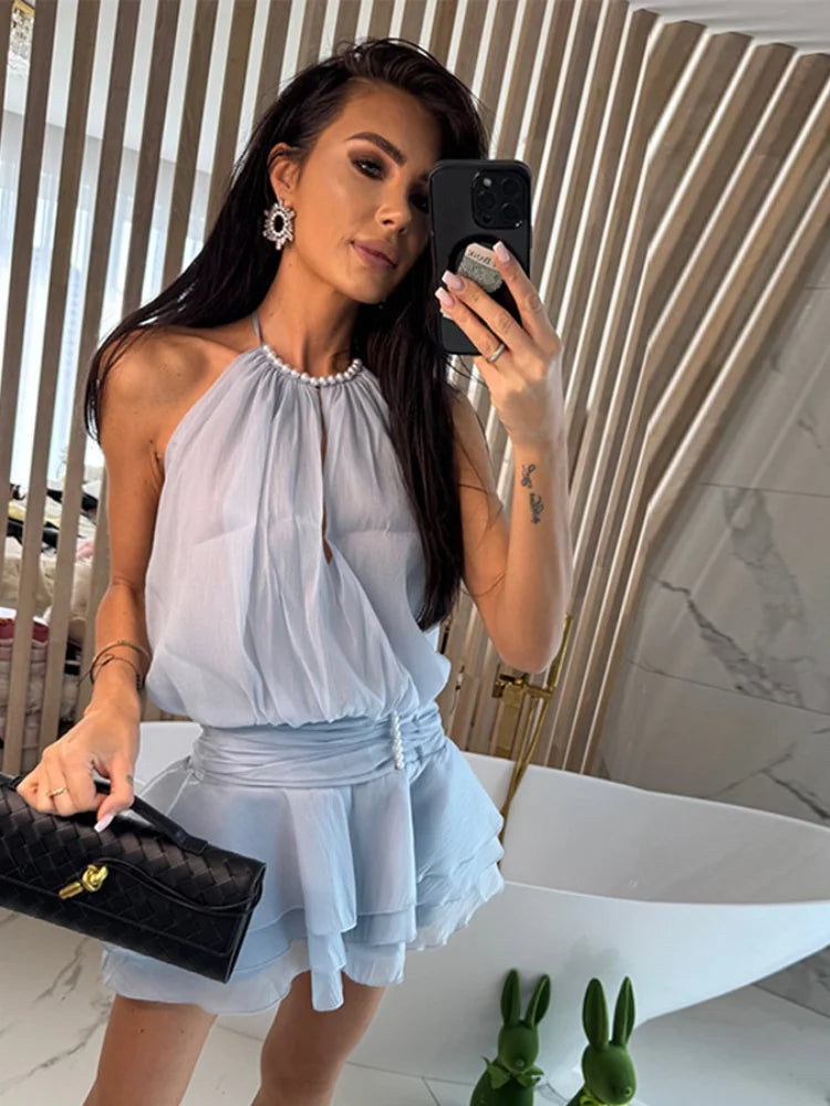 A woman taking a mirror selfie wearing a light blue ruffled chiffon mini dress with a halter neck and pearl detail in an indoor setting.