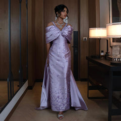 A woman wearing an elegant lilac off-shoulder mermaid gown with a flowing cape overskirt.