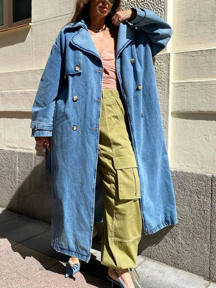 A person wearing a blue denim long trench coat with a belt, paired with green pants and a pink top.