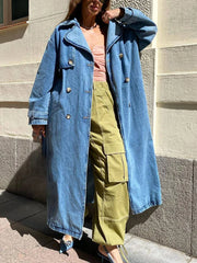 A person wearing a blue denim long trench coat with a belt, paired with green pants and a pink top.
