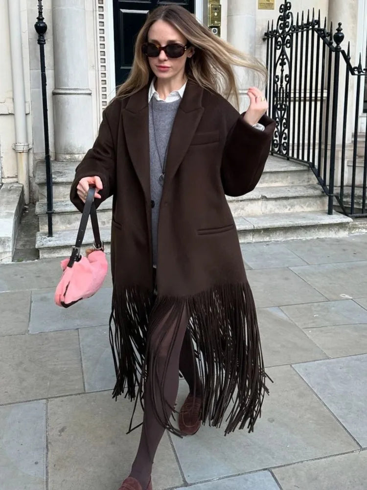 Walking moment highlighting the long brown fringe swinging from the trench hem, styled with sheer tights and boots for an evening look.