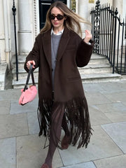 Walking moment highlighting the long brown fringe swinging from the trench hem, styled with sheer tights and boots for an evening look.