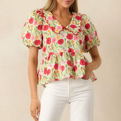 Woman wearing a short sleeve floral blouse with loose airy fit for summer with white pants on a beige background