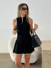 Woman in a black sleeveless dress holding a black handbag indoors.