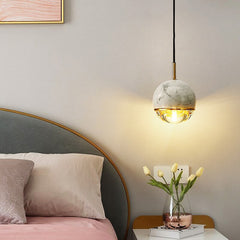 A  modern pendant light with marble and copper finishes,  suspended from the ceiling. Hanging beside a bed side table.