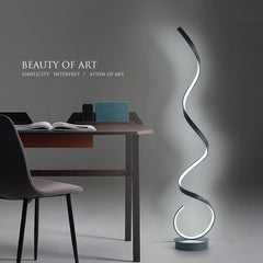 A modern black floating LED floor lamp with an illuminated golden spiral design, placed in a room with a dark wall and a study table and chair next to it.