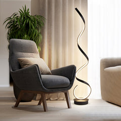 A modern black floating LED floor lamp with an illuminated golden spiral design, placed in a room with a light gray  wall and a sofa chair next to it.