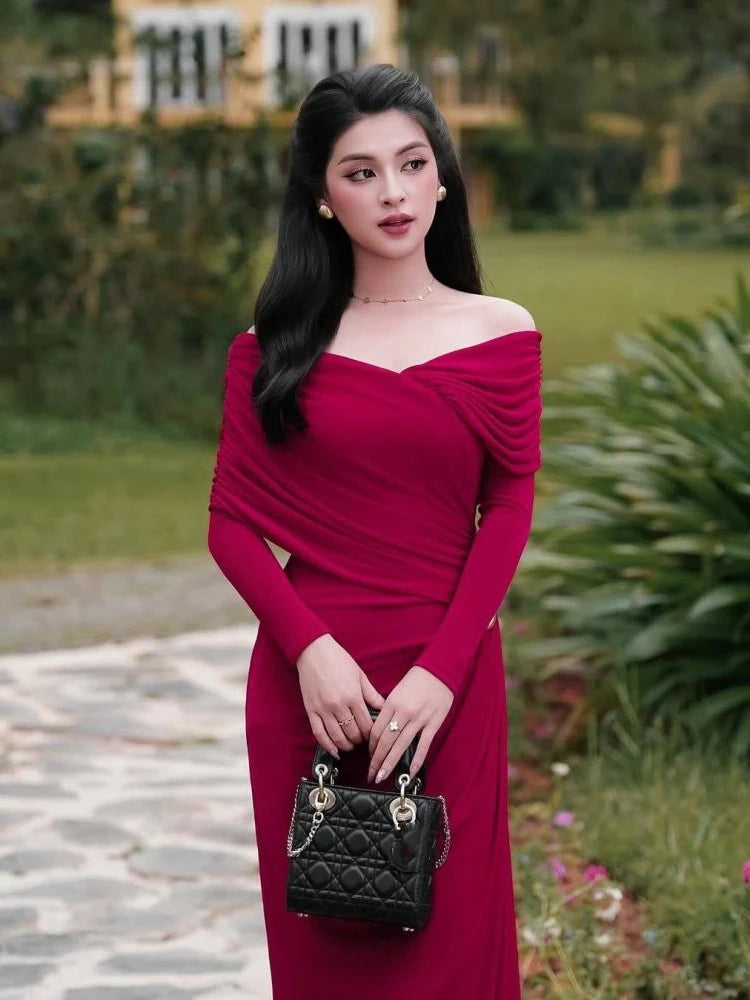 A woman in a folded, detailed, long-sleeved red party dress is shown on a stone path holding a black handbag outdoors.
