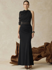 Model wearing a black one-shoulder top with a sheer sleeve and matching maxi skirt, styled with gold earrings in a studio setting.