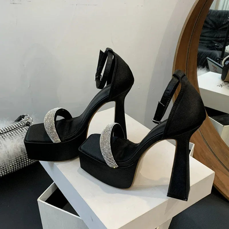 Black high-heeled shoes with rhinestone straps on a white platform, placed on a white surface.