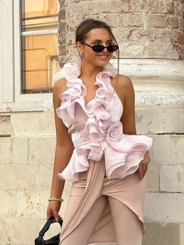 Woman wearing a pink ruffled top and beige pants standing against a stone wall.