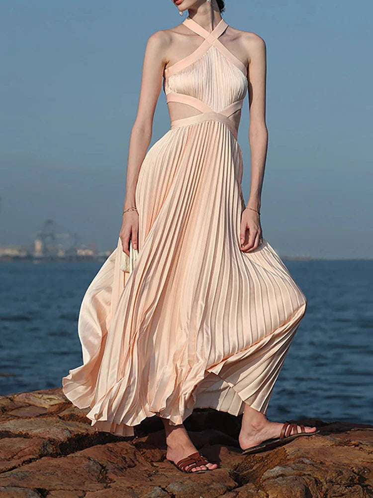 A woman wearing a pink halter pleated gown, flowing by the ocean, standing on rocks by the ocean.