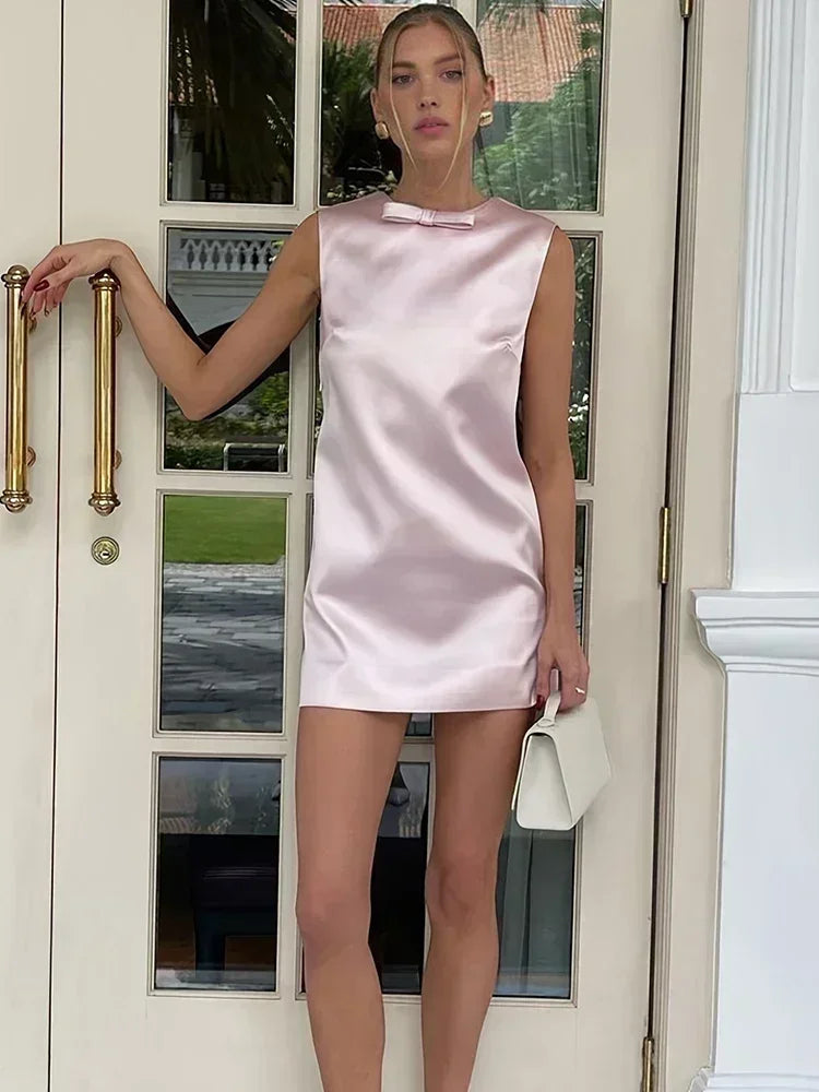 Side-back angle showing the open-back neckline and sleeveless cut of a pink satin mini dress styled with a small shoulder bag.