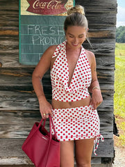 Woman in a polka dot halter crop top with coordinating mini skirt set holding a pink bag, standing in front of a wooden building with a Coca-Cola sign.