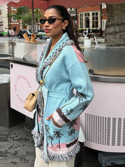 Street-style shot of a woman in a light blue printed long cardigan with tassel hem, sunglasses, and a crossbody bag near a kiosk.