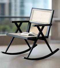 A black Scandinavian retro style rocking lazy getaway chair with a rattan weave pattern on the seat and backrest, a book on the lap, and a view of a cityscape through the frame.
