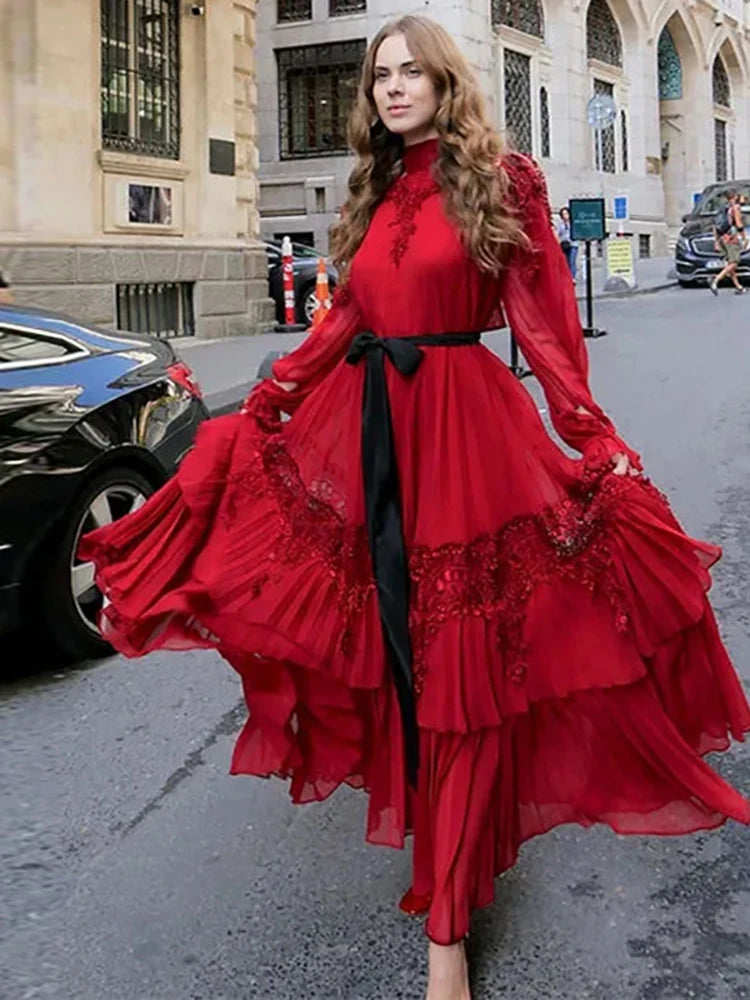 Woman in a red lace evening gown with long sleeves and a waist belt standing on a street.