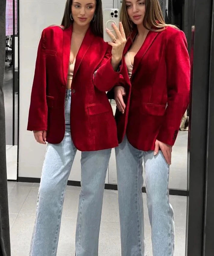 Two women wearing red single-button blazers with turn-down collars and blue jeans, taking a mirror selfie.