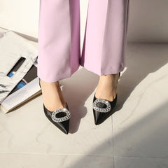 A pair of  black  stiletto pointed rhinestone buckle satin pump shoes with thin heels, displayed  by a model  wearing  a light  violet  pant.