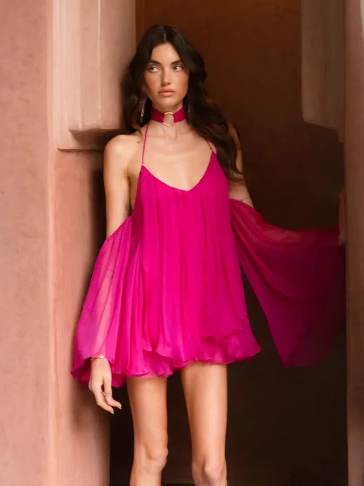 Woman wearing a rose red  halter mini dress with open back and flowy sleeves against a warm-toned wall.