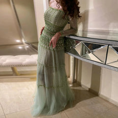 Women in a Sage green off-shoulder evening dress with beaded long sleeves, standing in an elegant room.