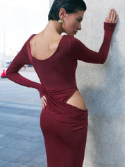 A woman wearing a sculpted long-sleeve evening gown for formal autumn events with a cut-out design, standing against a light-coloured wall.