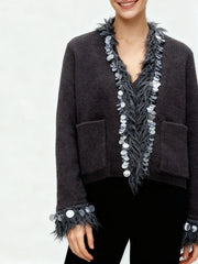 Relaxed knit jacket with sequin and tassel trim styled casually over dark trousers in a minimalist studio setting.
