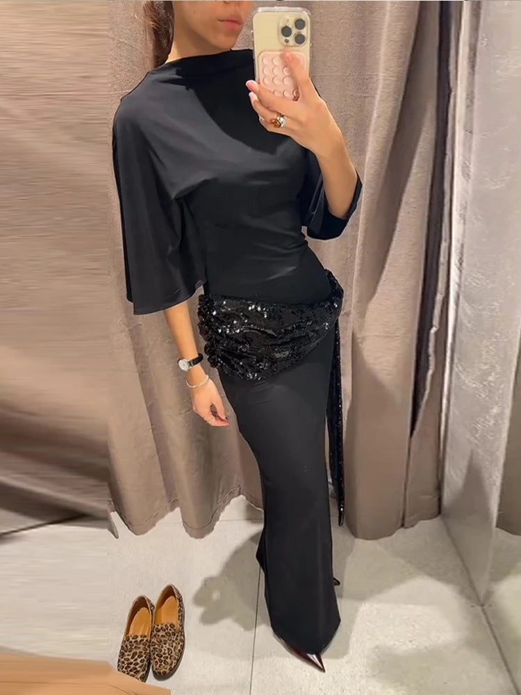 Woman wearing a black mermaid maxi dress with sequin waist, photographed indoors against soft curtains, highlighting the sculpted silhouette.