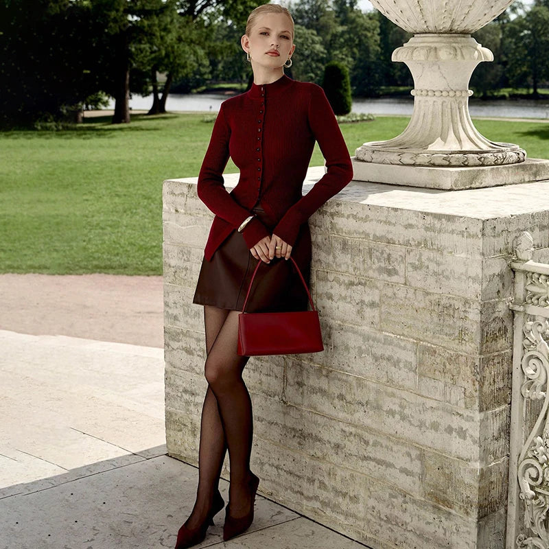 Woman in a  single breasted autumn winter sweater in vibrant red for juniors and brown skirt holding a red handbag against a stone wall outdoors.