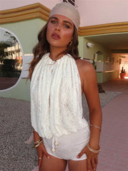 A woman wearing a backless white lace romper and tie halter straps with a beige headband, standing in front of a building.