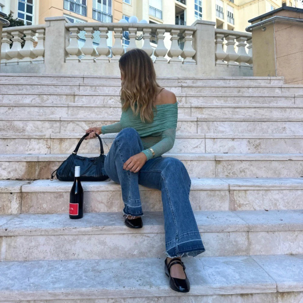 Woman, Slim fit  teal long sleeve one-shoulder top styled for spring summer sitting on stone steps with a bottle of wine and a handbag