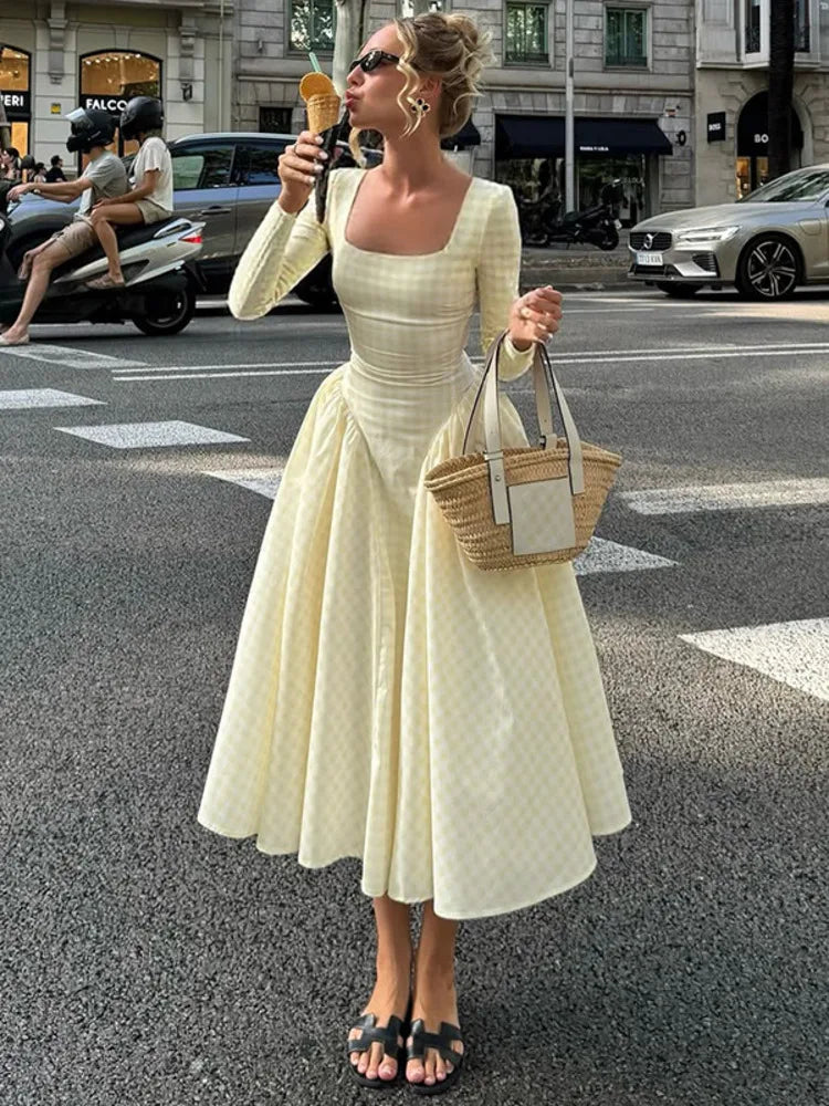 Woman in a square-neck fall dress with long sleeves and pleated skirt holding a wicker bag and ice cream cone on a city street.