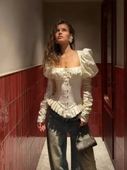 Studio shot of a woman wearing an ivory square neck corset blouse with puff sleeves, styled with black leather-look pants on a grey backdrop.