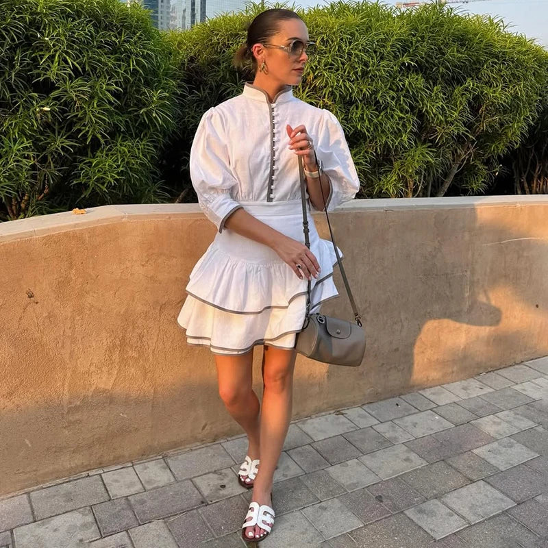 Woman in a stand collar high waist ruffle dress in white for commuting chic holding a gray handbag against a green hedge background