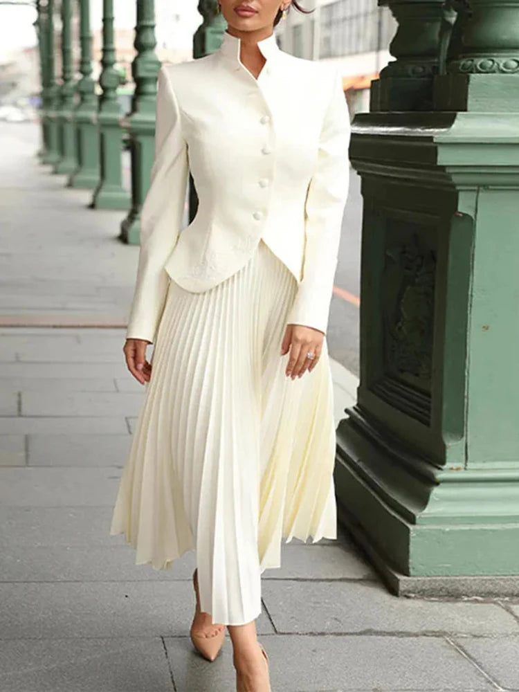 Model wearing a light-toned stand collar top and pleated high-waist skirt set, styled outdoors in a formal setting.