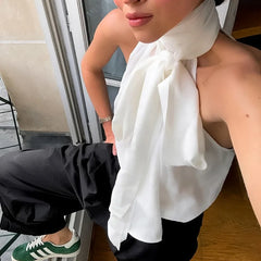 A person wearing a statement bow party sleeveless blouse in white is shown with satin trousers and black pants sitting on a bench.