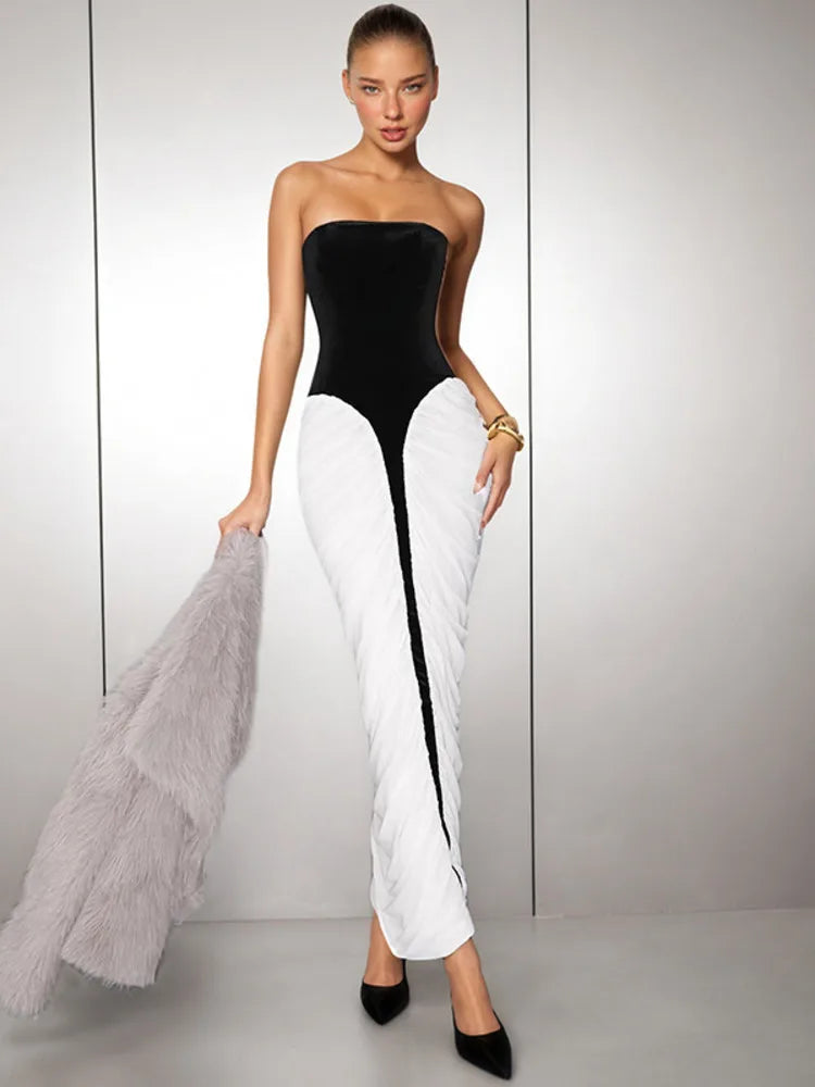 Woman wearing a strapless black and white maxi dress styled for night events, holding a grey fur coat against a light grey background.