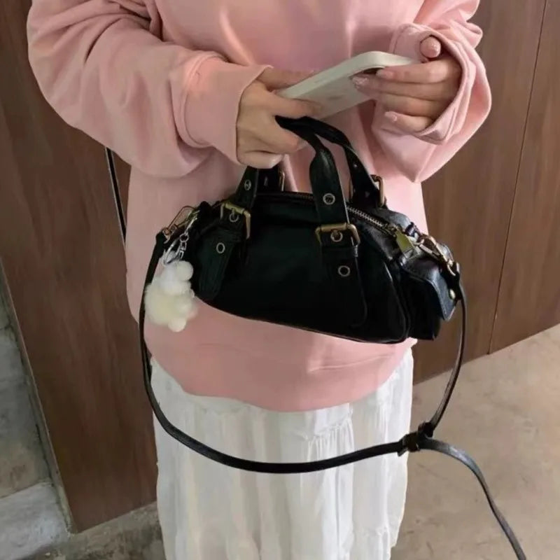 Person holding a black leather crossbody satchel styled for high street fashion with a white flower charm against a wooden background